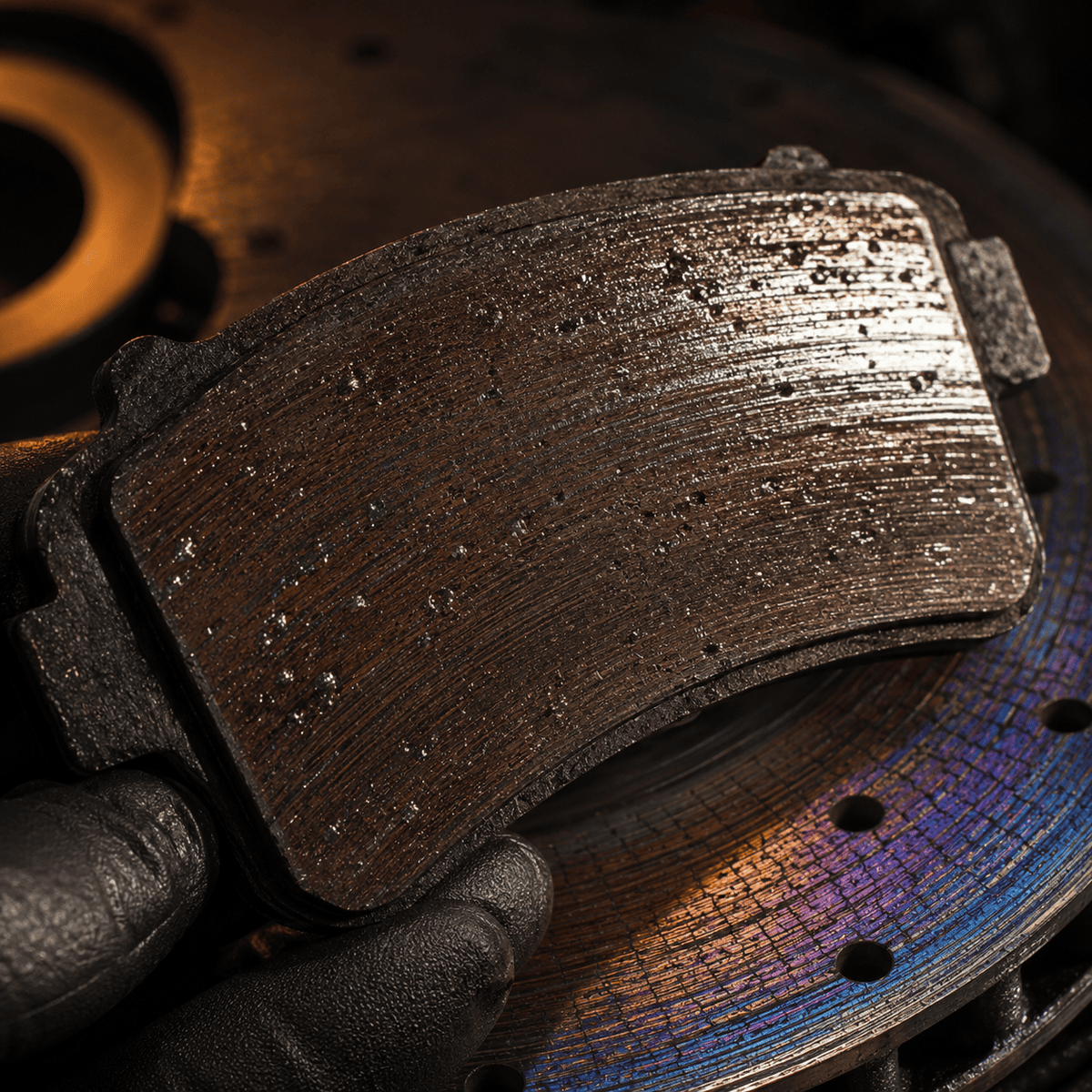 Macro close-up of a used motorsport brake pad with a glazed, vitrified friction surface held above a heat-discoloured rotor — both surfaces showing clear signs of being pushed beyond the compound's operating window.